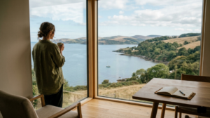 A person looks out a large window at a peaceful bay, standing beside a wooden table with an open journal.